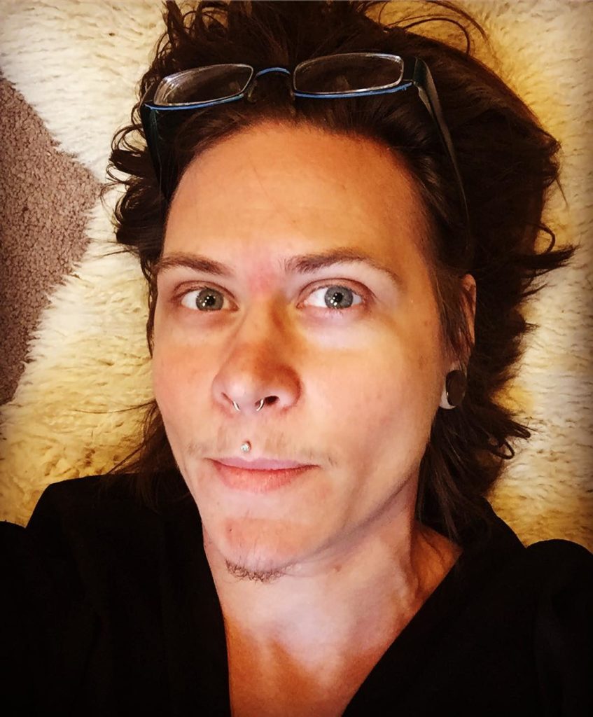Headshot of Creature Karin Webb. Pierced septum and medusa, glasses on top of forehead. Medium length light brown hair, light chin hairs, faint sparse mustache, blue eyes.